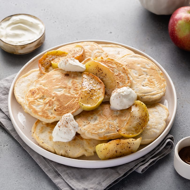 A plate of pancakes with caramelized apple slices and dollops of whipped cream, sprinkled with spices.
