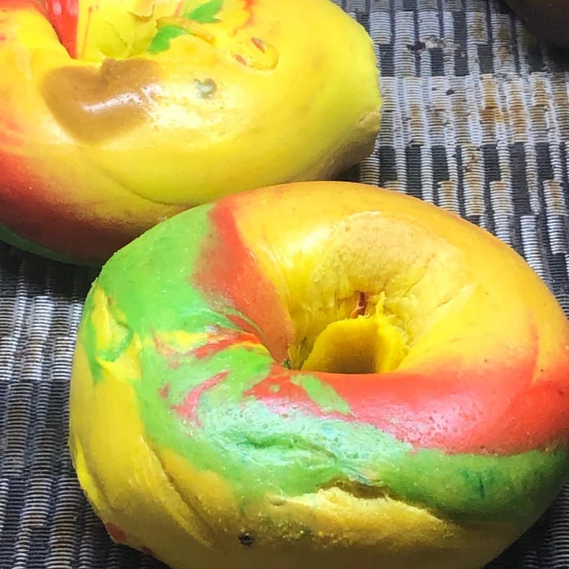 Two vibrant rainbow bagels, swirled with red, green, and yellow, rest on a dark patterned surface.