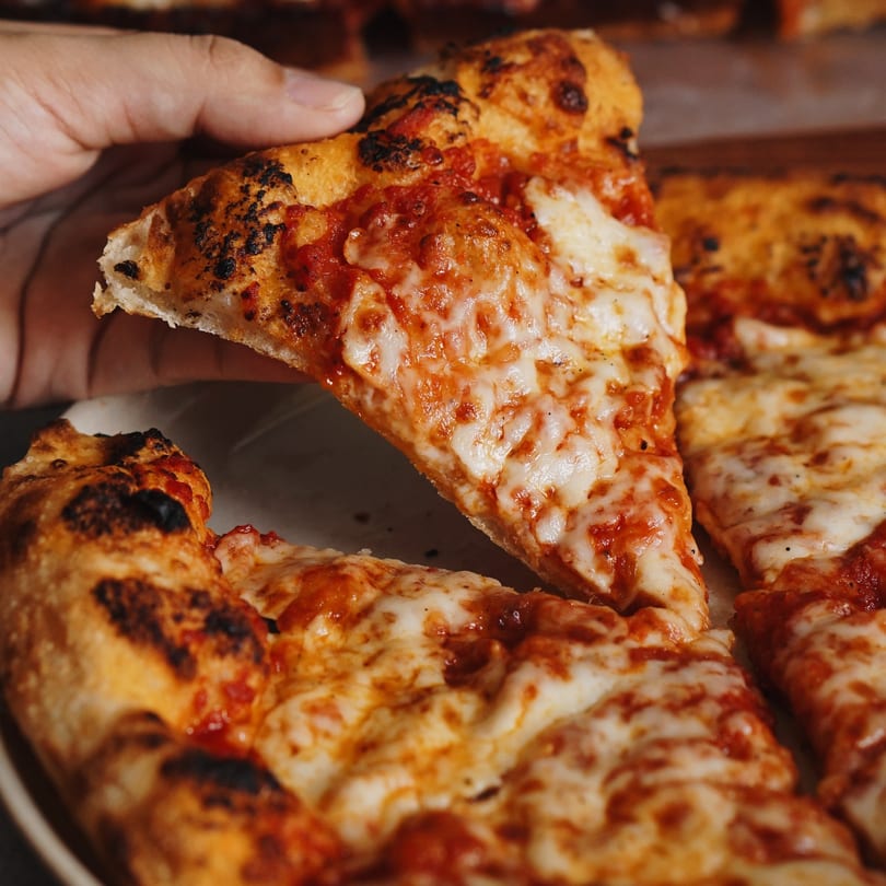 A hand lifts a slice of cheesy pizza with a crispy crust from a whole pizza.