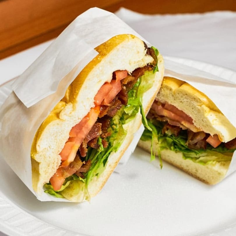 A Tribeca Bagels BLT sandwich, cut in half, wrapped in white paper on a white plate.
