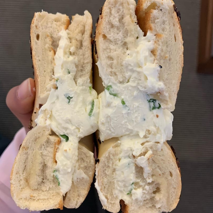 A Tribeca Bagels bagel, sliced in half, overflowing with cream cheese and green herbs.