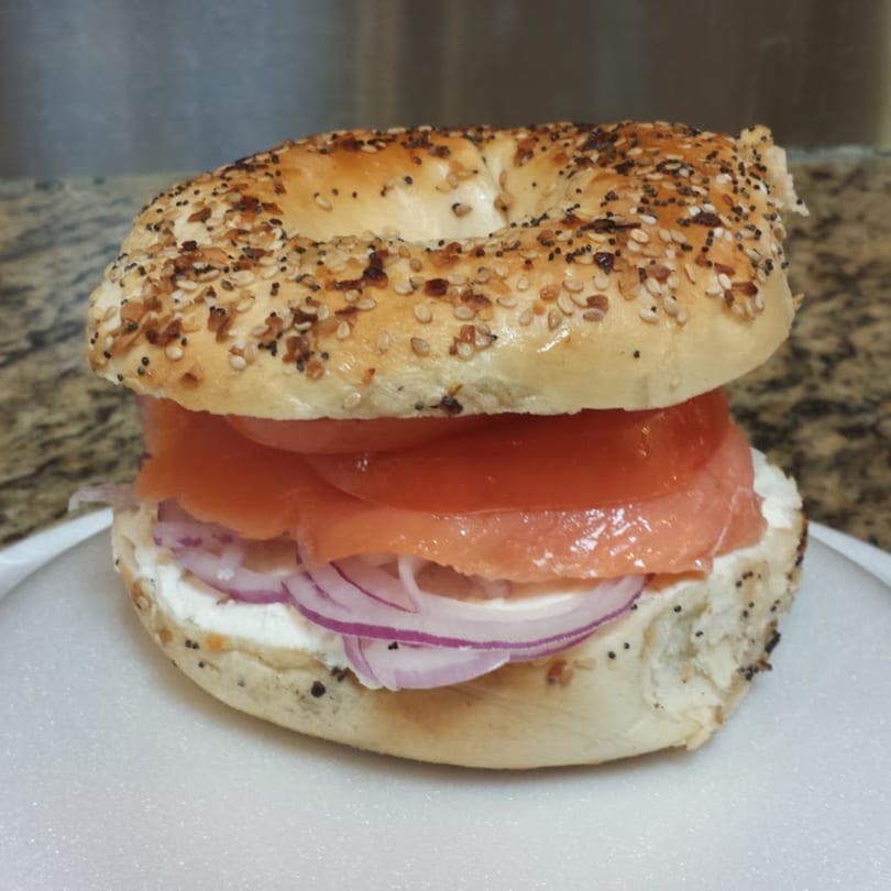 Everything bagel with cream cheese, smoked salmon, sliced tomatoes, and red onions on a white plate.